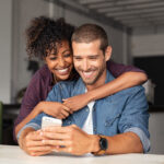 Smiling,Young,Couple,Embracing,While,Looking,At,Smartphone.,Multiethnic,Couple